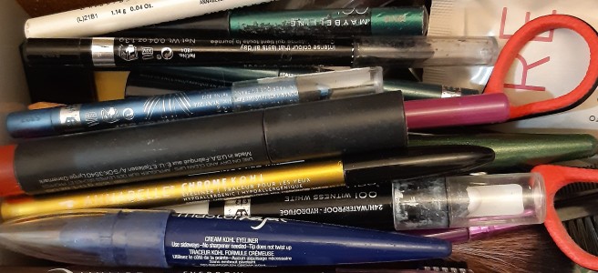 A close overhead photo of eye liner pencils, lipstick tubes, and make-up brushes thrown together in a plastic box.